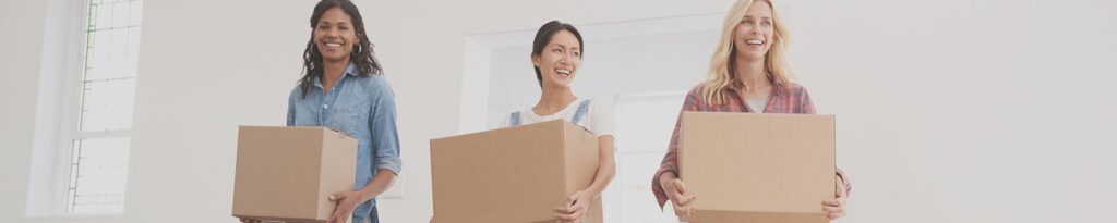 Three smiling women carrying moving boxes into a shared home, illustrating HMO mortgages for UK property investors.